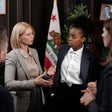 A woman in a gray blazer and beige blouse gestures while speaking to another woman in a black blazer and white shirt, standing in an indoor office-like setting with a California flag and dark wood furniture in the background.