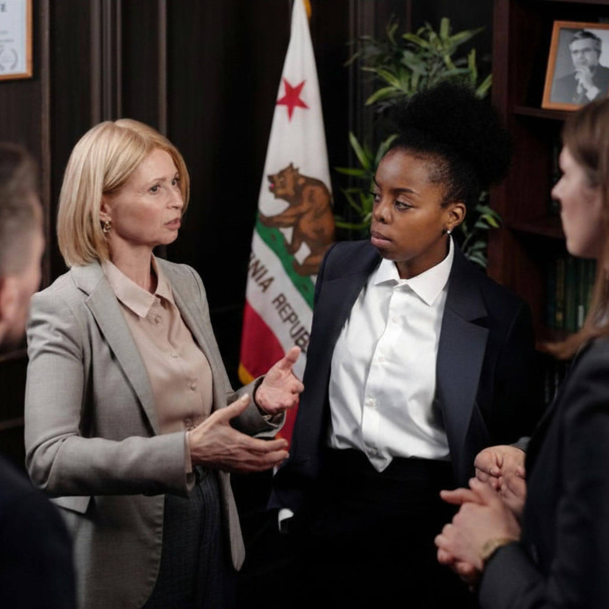 A woman in a gray blazer and beige blouse gestures while speaking to another woman in a black blazer and white shirt, standing in an indoor office-like setting with a California flag and dark wood furniture in the background.